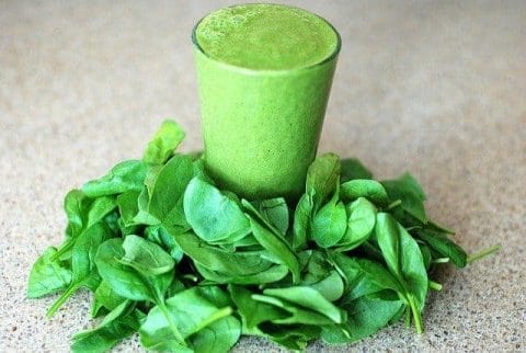 Un vaso con un batido verde, con hojas de espinacas alrededor