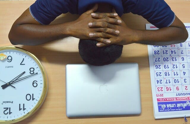  Un hombre cansado mantiene la cabeza sobre su escritorio, junto a su portátil, su calendario y su reloj